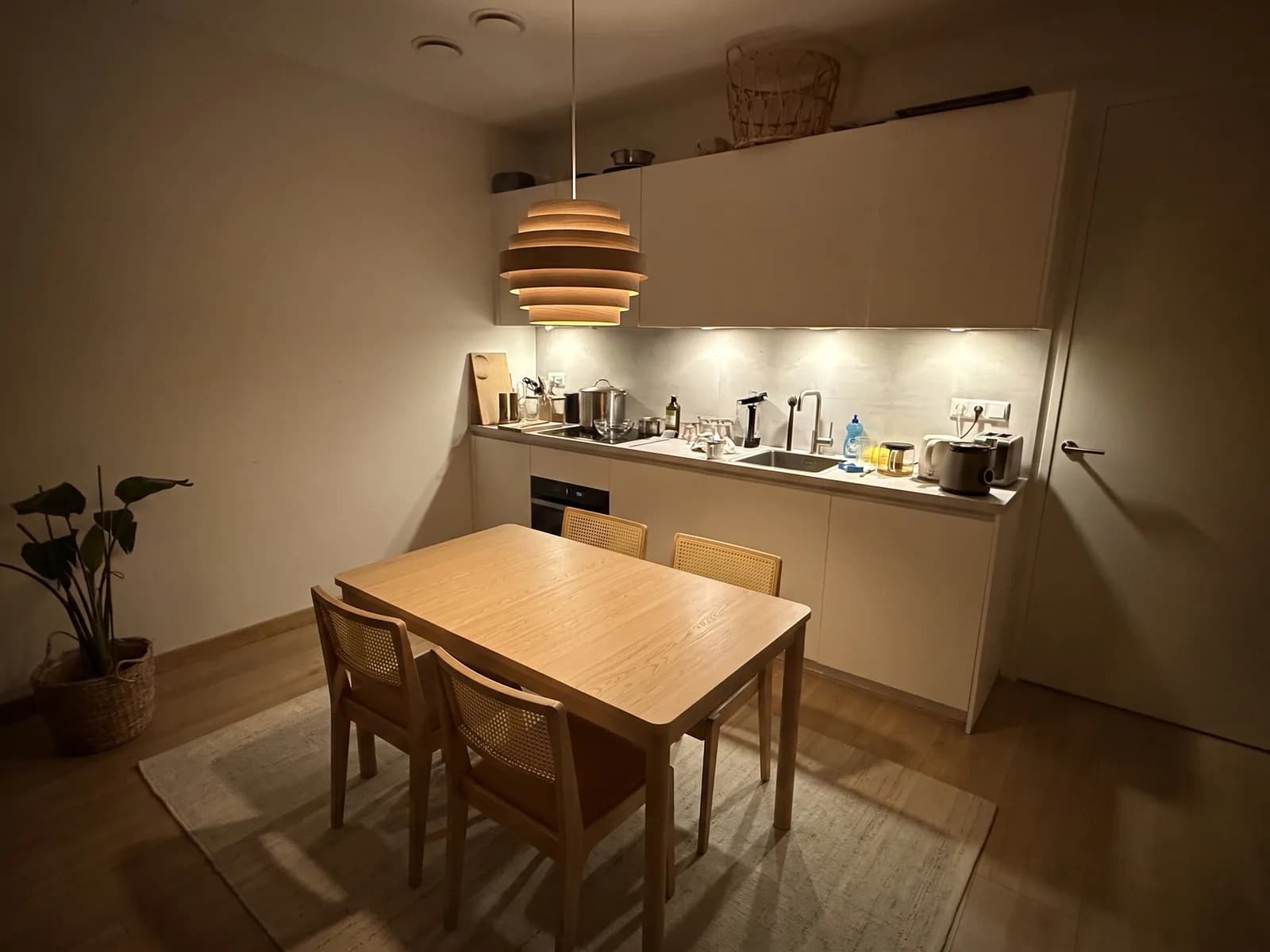 The same kitchen with an oak TONSTAD table, four rattan STOCKHOLM chairs, a beige rug and a wooden pendant lamp
